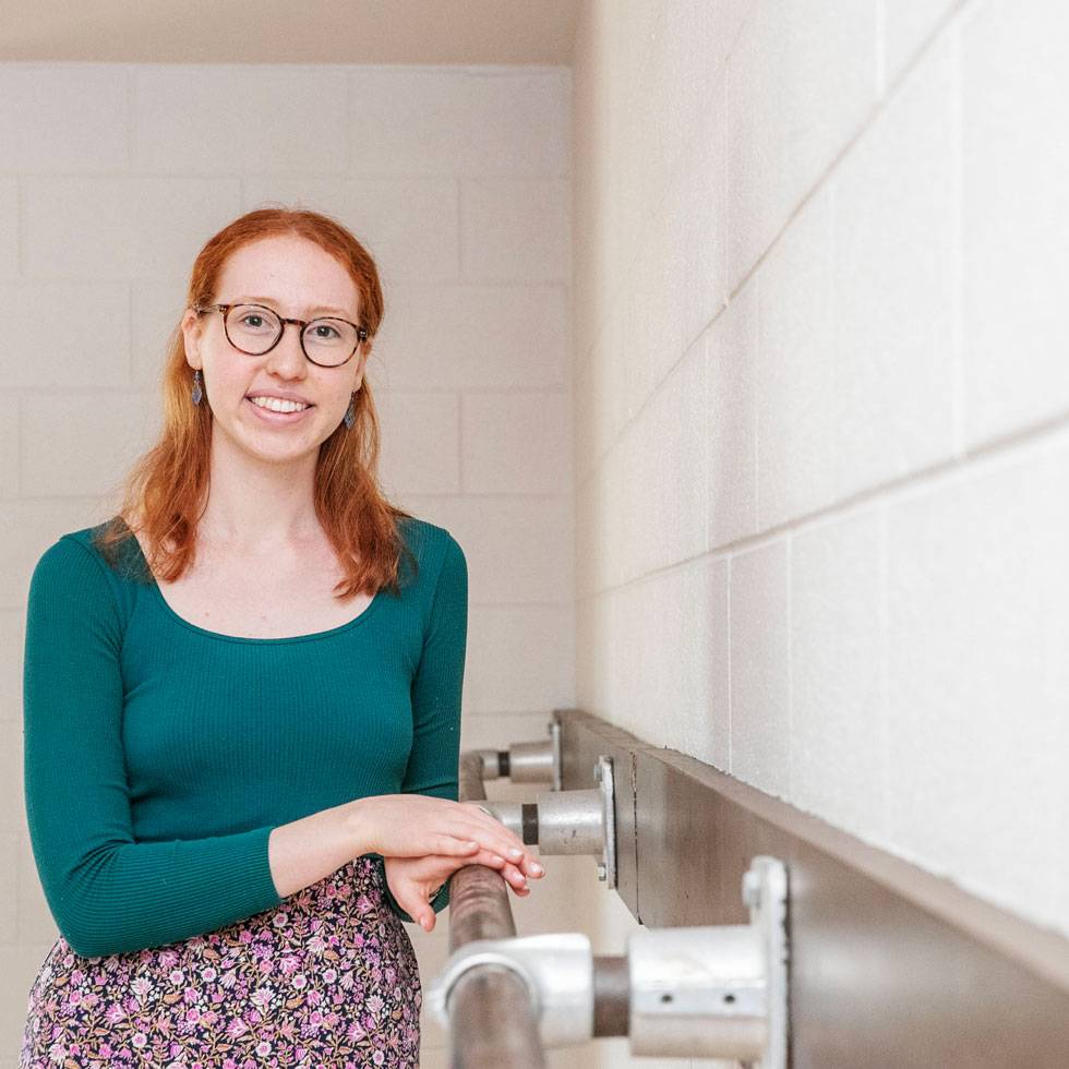 Liz Bracht stands in a green shirt and floral skirt hold her hands on a metal dance bar with white walls all around.
