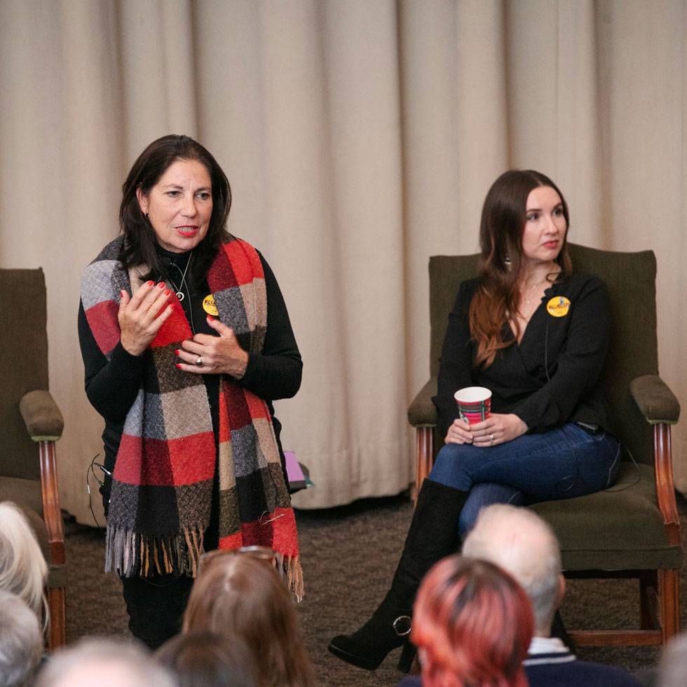 A women speaks to an audience while another is seated in a chair behind her. The foreground shows the backs of the audiences heads.