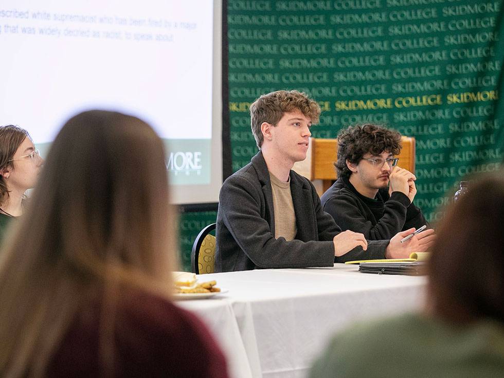 Two students sit in a discussion group with a screen and Skidmore banner in the background.