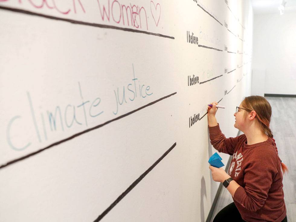 A student crouches down while writing on a white wall with a blank space to fill in the blank after "I believe..."