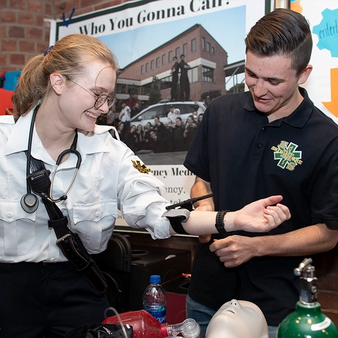 Two Skidmore students in EMS uniforms share a laugh while practicing blood pressure measurement at a club fair.