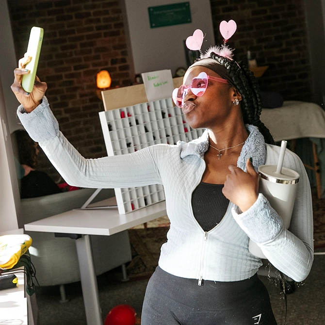 A student wearing heart-shaped glasses and holding a tumbler poses for a selfie at a festive, student-led event.