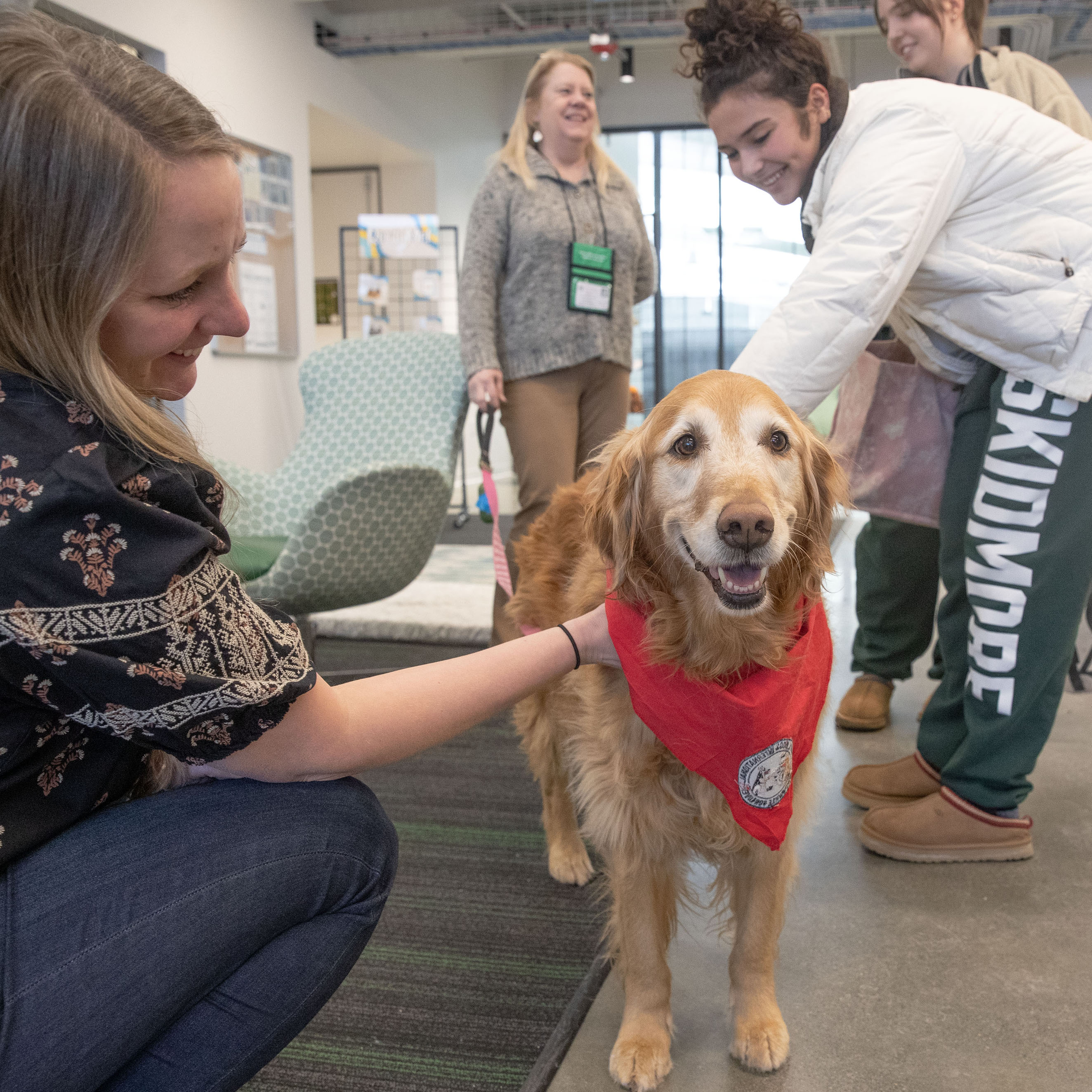 Students gather around and pet a golden retriever wearing a red bandana during a therapy dog event on campus.