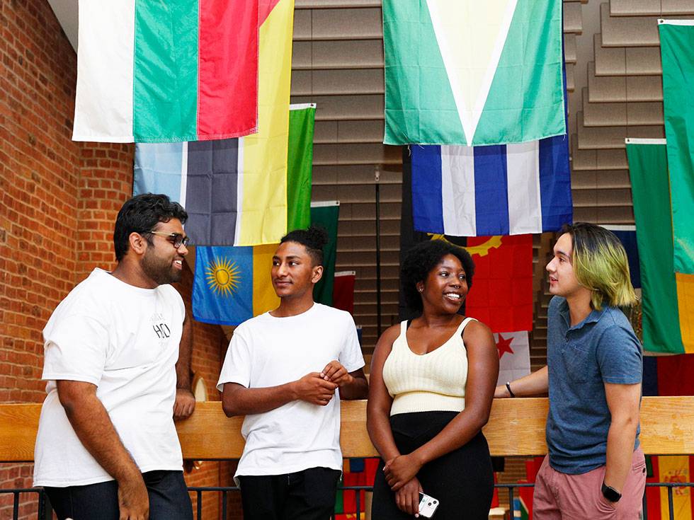 Four international students stand in a line talking with the various flags in the background hanging from the ceiling of Case Center.