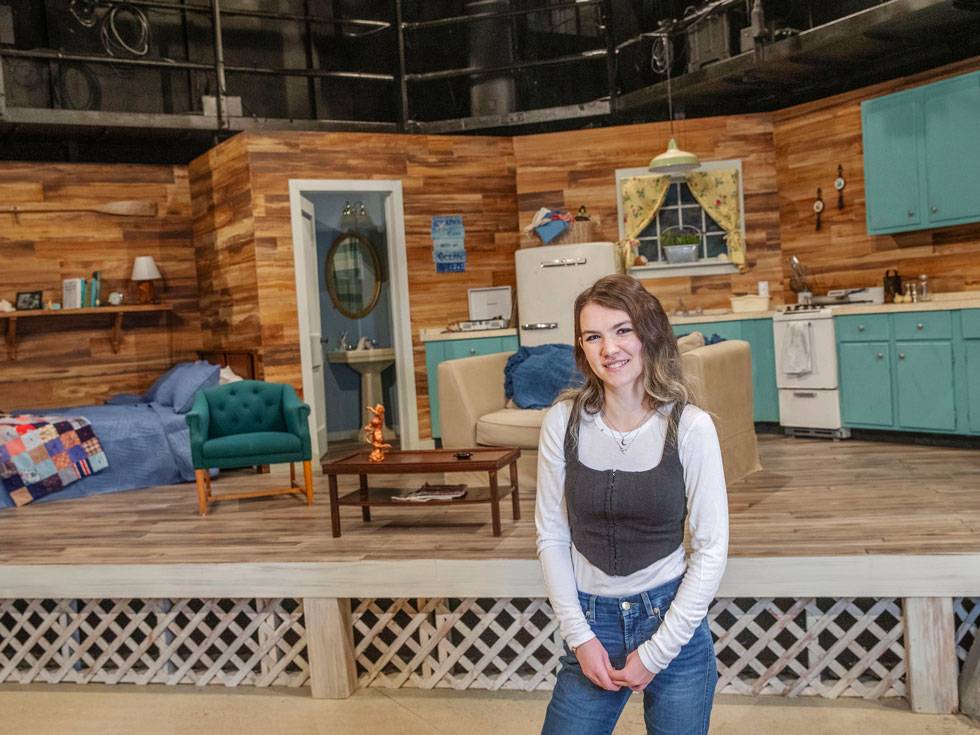 A young woman stands while smiling in front of a detailed stage set featuring a rustic wooden interior, including a bed, vintage refrigerator, small kitchen area, and living room furniture.