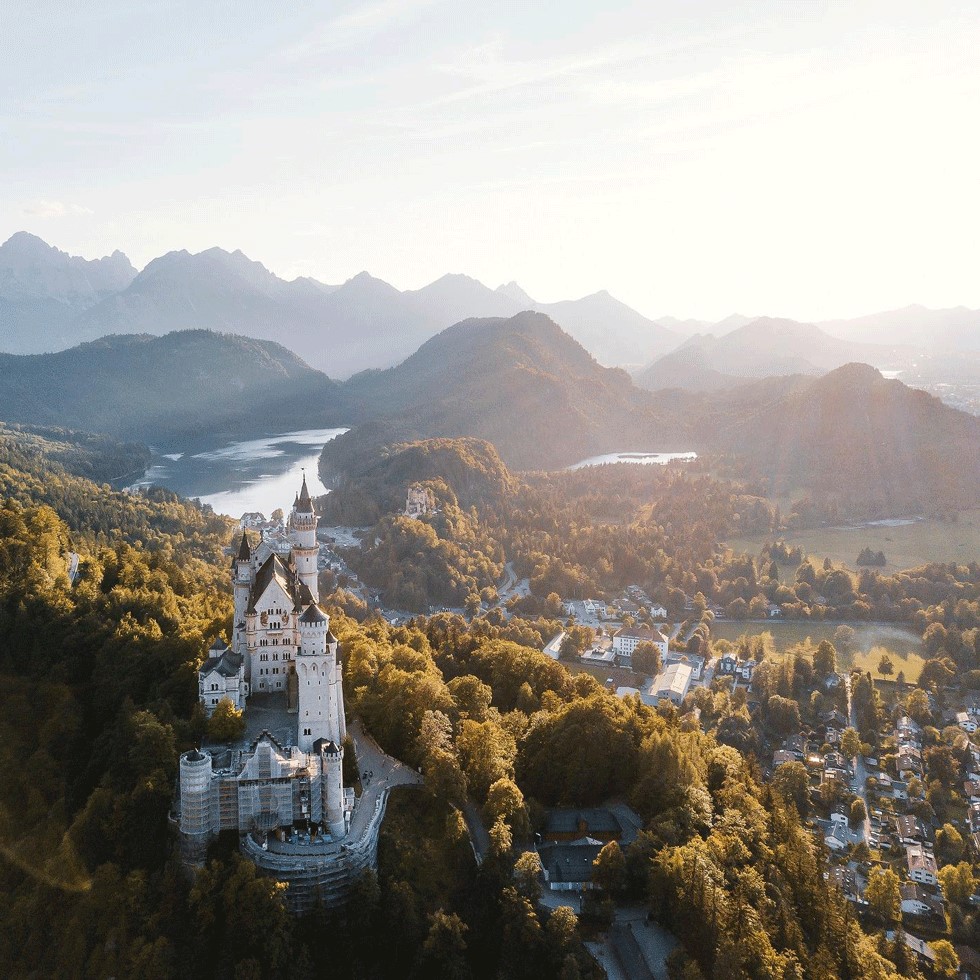 German castles sits on a hillside with town below and mountains and lakes in the background.