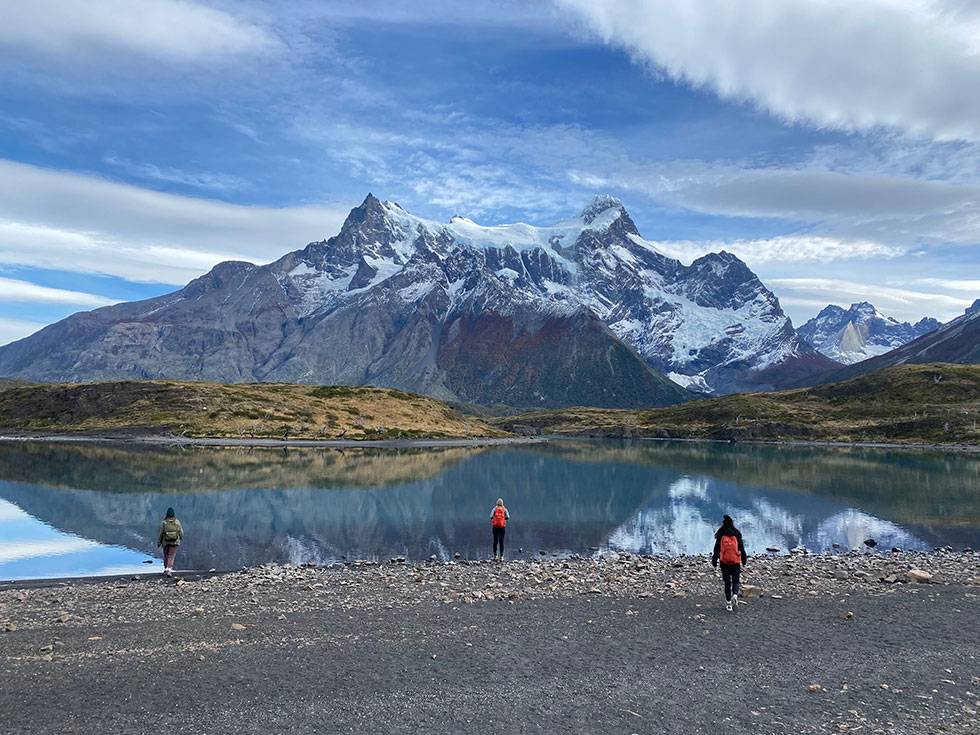 Three people walk in Torres del Paine with a reflective lake and snow capped mountains in the background