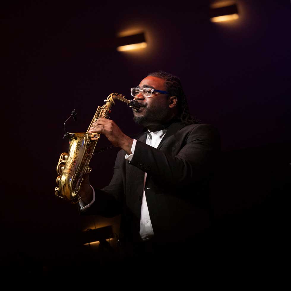 A saxophonist performs onstage in Zankel Music Center against a dark background with warm stage lighting.
